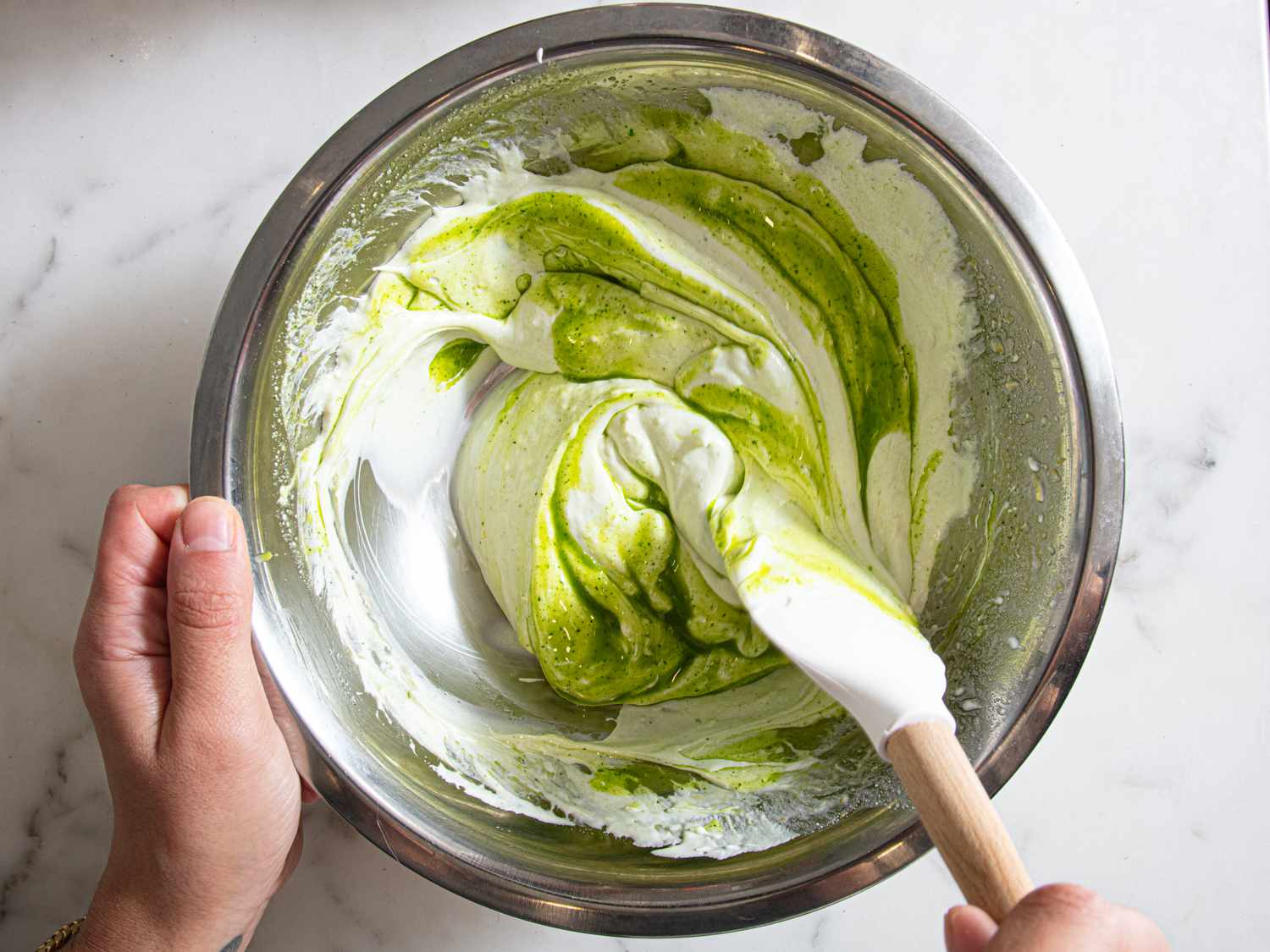 Hands stirring a mixture with a spatula in a stainless steel bowl