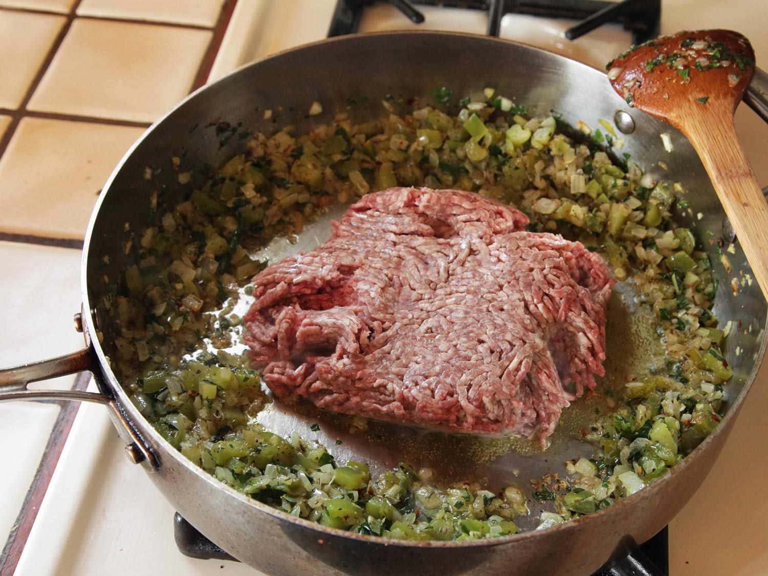 The onion, green pepper, and aromatics have been pushed to the periphery of the pan to make room for a large mound of raw ground beef.