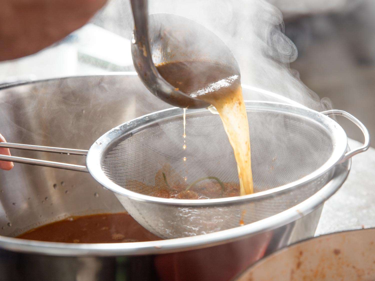 The finished lobster stock being ladled through a fine-mesh strainer and into a large mixing bowl.