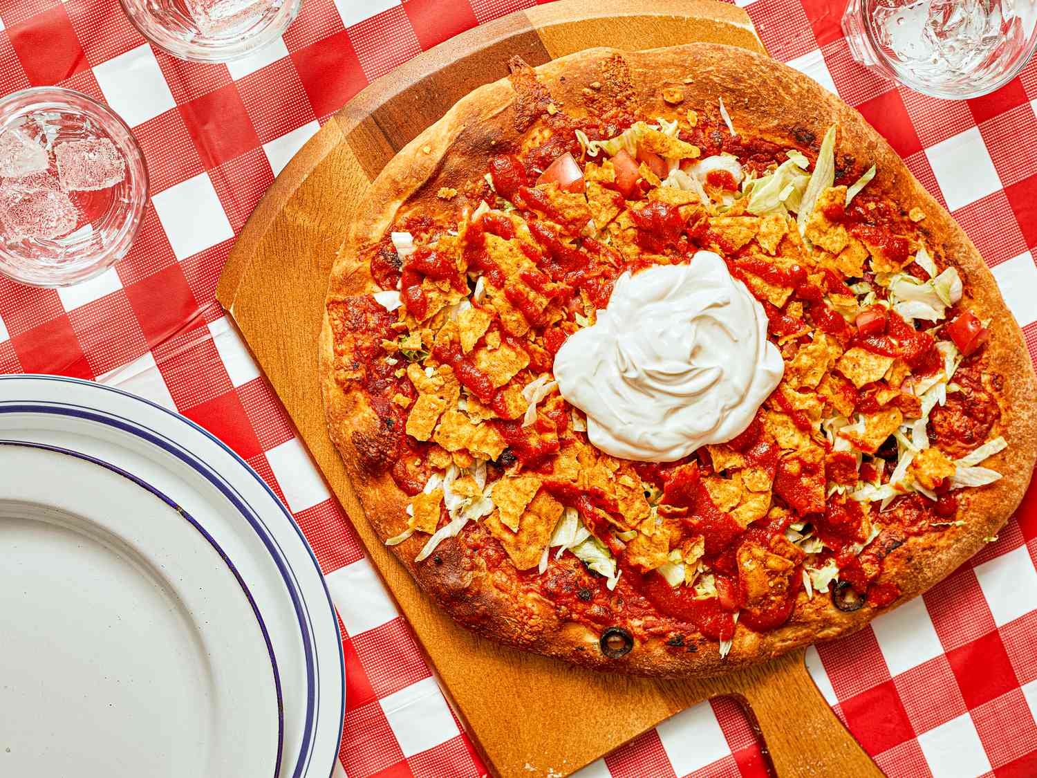 Taco pizza with sour cream on a wooden board, set on a red-checkered tablecloth, plates, and glasses nearby.