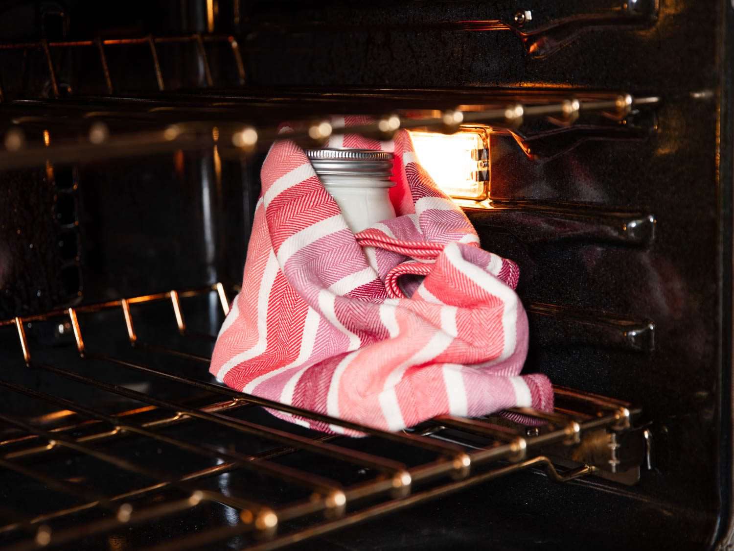 A mason jar of yogurt wrapped in a towel and placed in a turned-off oven with the light on.