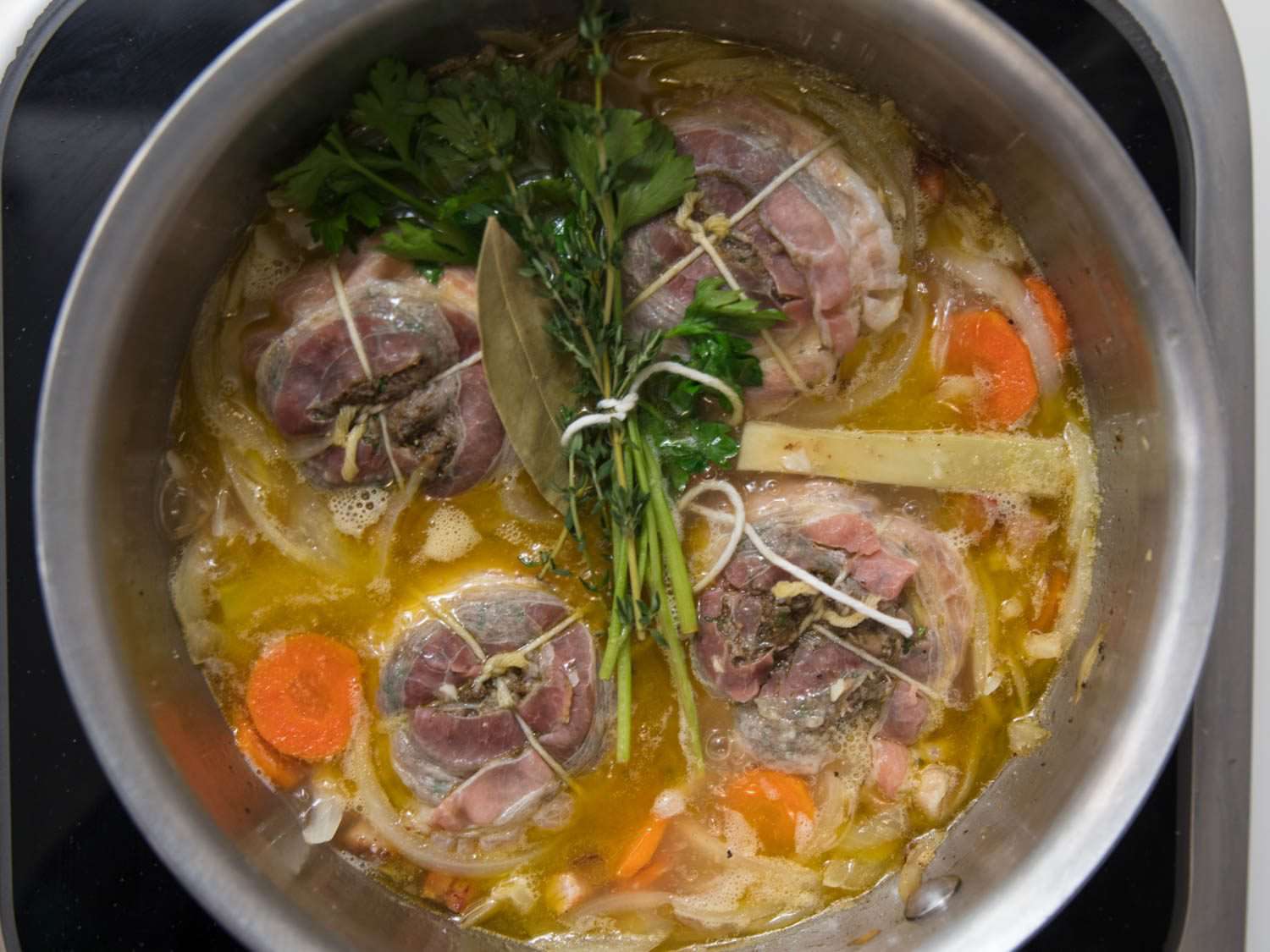Overhead view of stuffed, pancetta-wrapped artichokes, aromatics, and stock in a saucier pan.