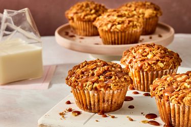 apple pie muffins on a white tray, with 3 muffins on a circular tray in the background. A clear glass of milk to the side on pink napkins. 
