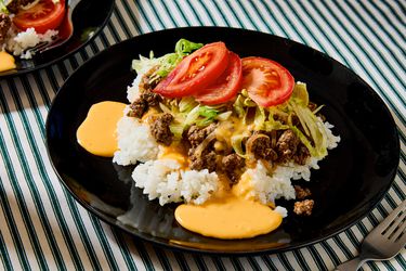 A plate of taco rice with beef, lettuce, and sliced tomatoes on top of white rice, served on a striped tablecloth
