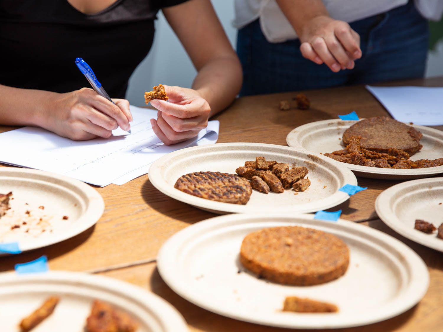 Tasters writing tasting notes in the background, with numbered paper plates with bare meat-replacement burger patties in the foreground.