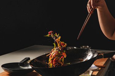 Colorful food suspended in air caused by a person tossing a wok while holding chopsticks in a dark kitchen