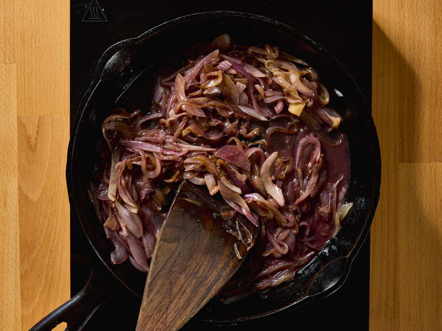 Caramelized onions cooking in a cast iron skillet on a stovetop with a wooden spatula