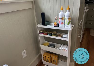 A white kitchen storage rack with olive oil, spices, napkins, and dried goods on its shelves