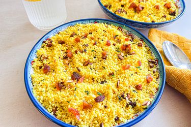 Bowls of couscous garnished with dried fruits and nuts on a table near a drink and a yellow napkin with spoon
