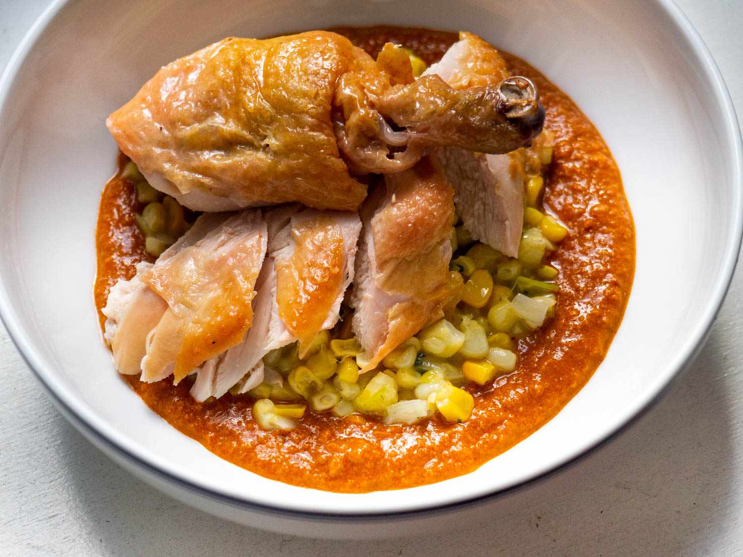 Closeup of a bowl containing sliced roast chicken pieces served atop a puddle of romesco crowned with a smattering of creamy corn.