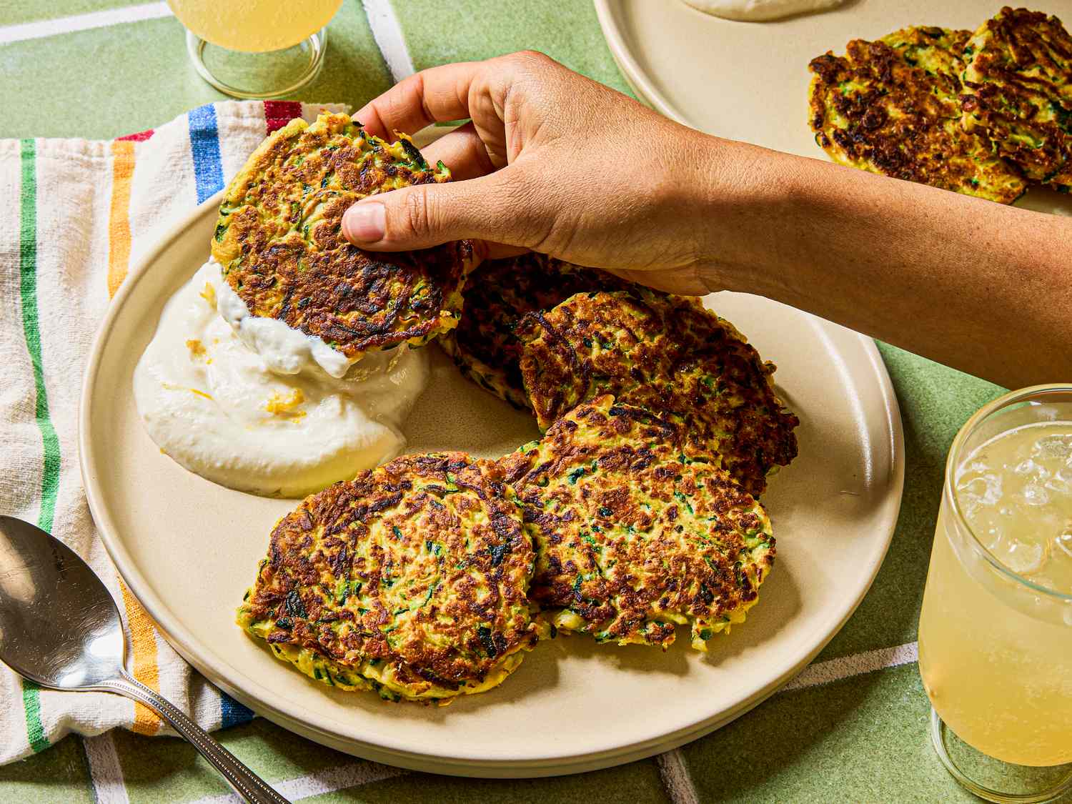 A hand placing a zucchini pancake on a plate with other pancakes and a dollop of cream