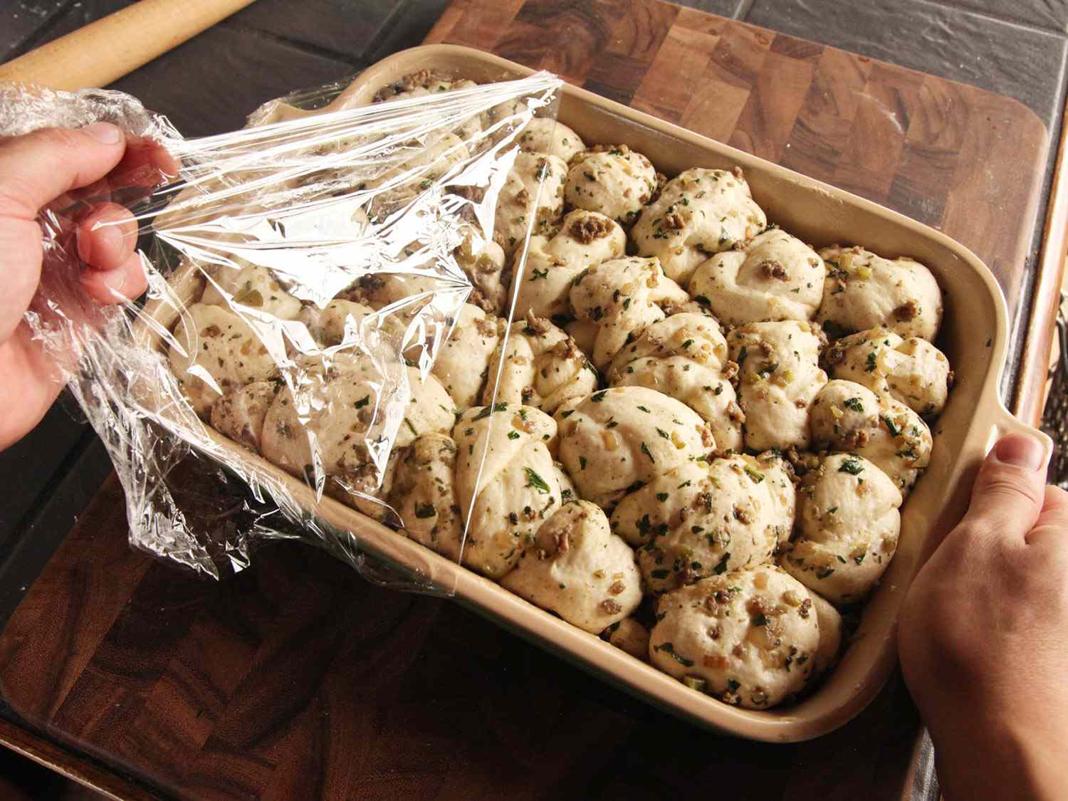 Pulling off a layer of plastic wrap over a baking dish of unbaked pull-apart stuffing rolls, roughly doubled in size after rising