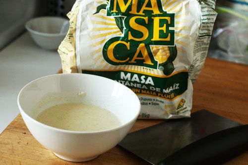 A bag of masa harina on a countertop. A white bowl containing a slurry of masa harina and water is placed in front of the bag.