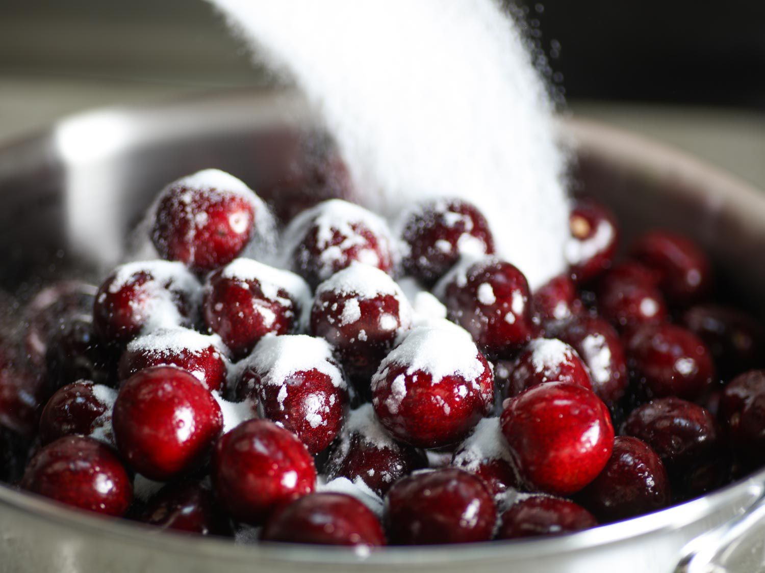 Adding sugar to a stainless steel skillet full of cherries.