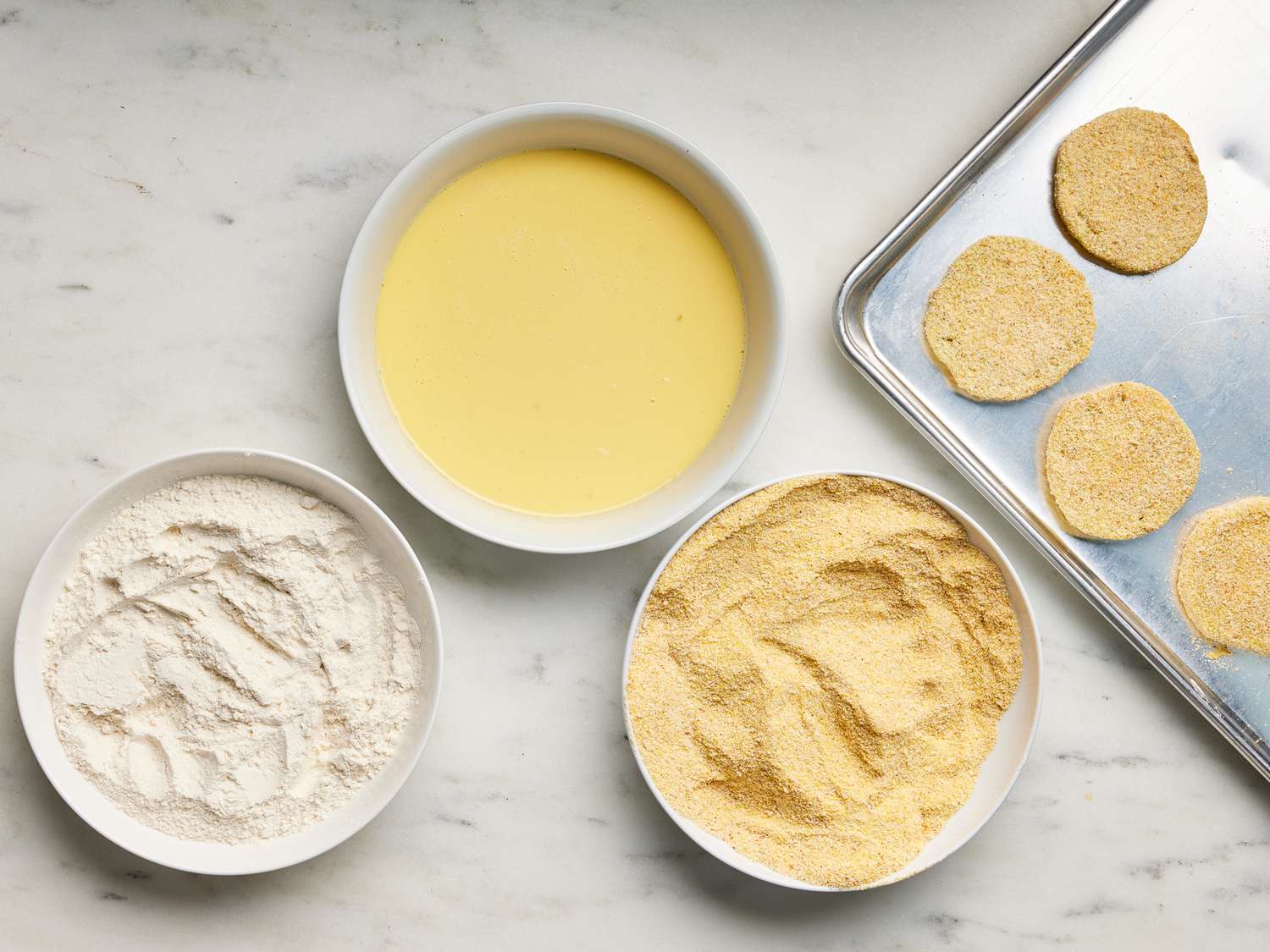 Cornmeal, egg and flour in 3 separate bowls with dipped and breaded tomatoes on a sheet pan next to them