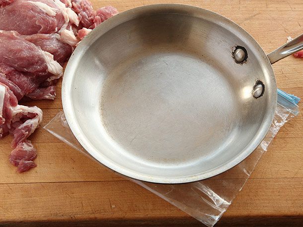 Heavy skillet resting on plastic zip top bag beside pile of sliced raw pork on wooden board