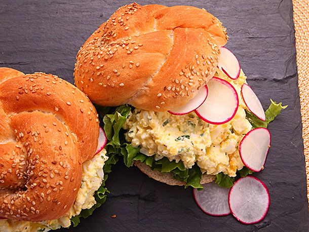 A pair of egg salad sandwiches made with kaiser rolls. The top of one roll is resting to one side of the sandwich and radish slices are cascading off the other.