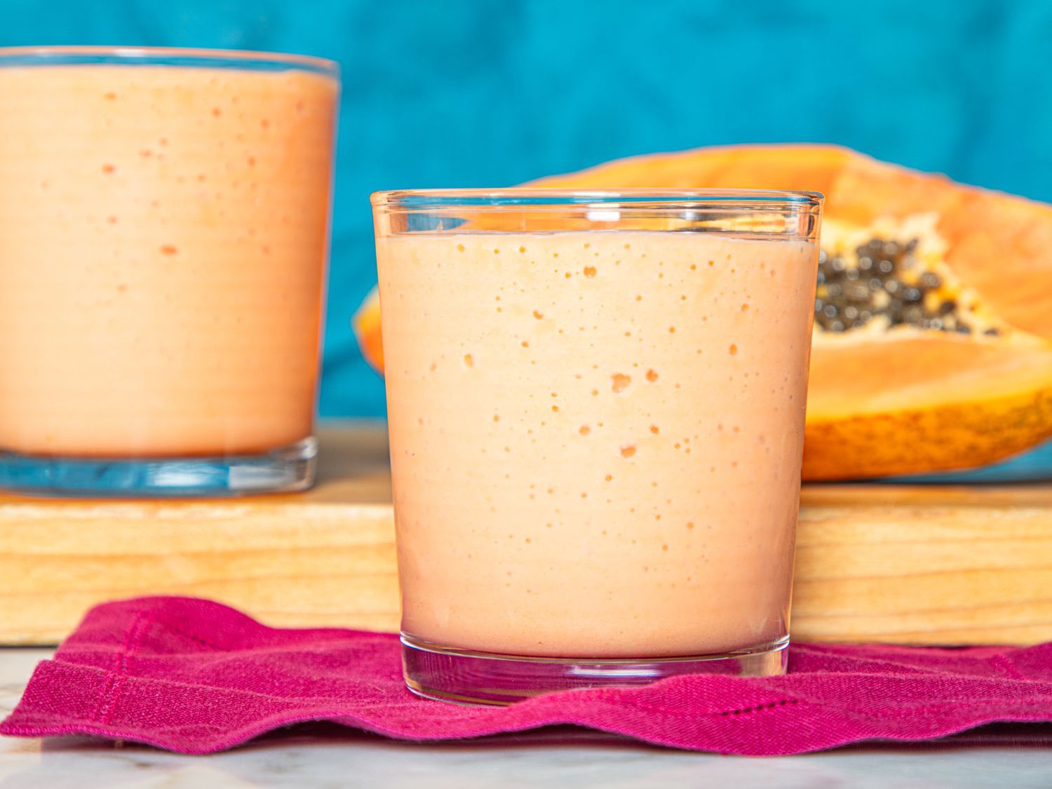 Two glasses of fruit smoothie on a pink napkin wooden board with papaya in background