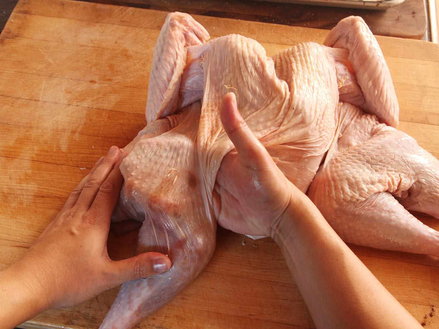 A hand reaches underneath the skin of a raw turkey to separate the skin from the flesh.