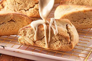 Maple scone being drizzled with glaze displayed on a cooling rack with other scones in the background