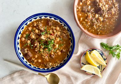 Bowl of Chobra Frik in a blue and white bowl, with small dish of lemons and cilantro on a white marble background