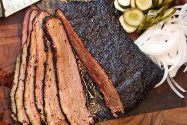 Cooked, sliced sous vide brisket on a cutting board with thinly sliced onions and pickles. 