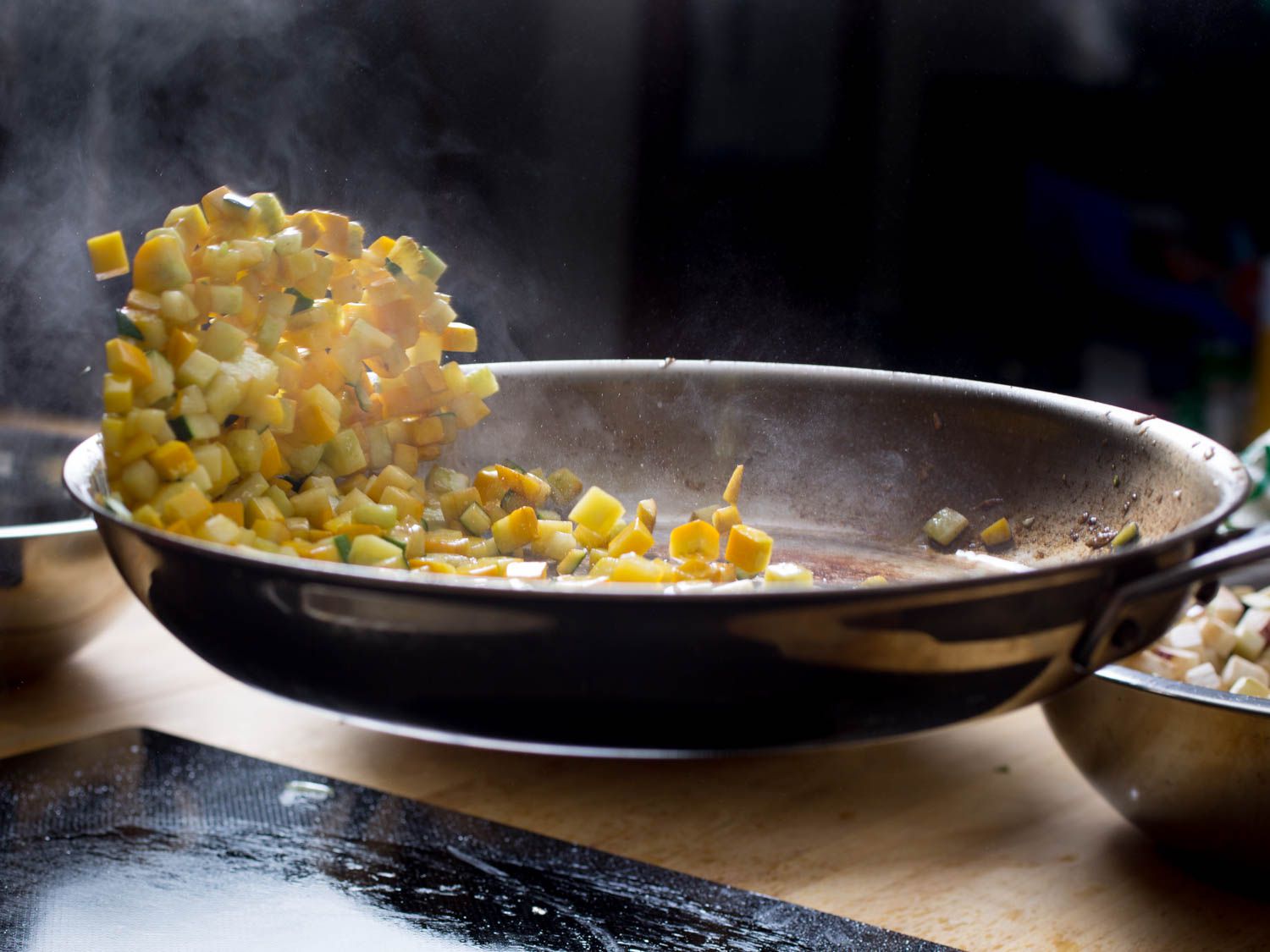 Diced squash being sautéed in a skillet. The author is tossing the vegetables in the pan by deftly flicking the pan so that the pieces of squash are launched into the air.