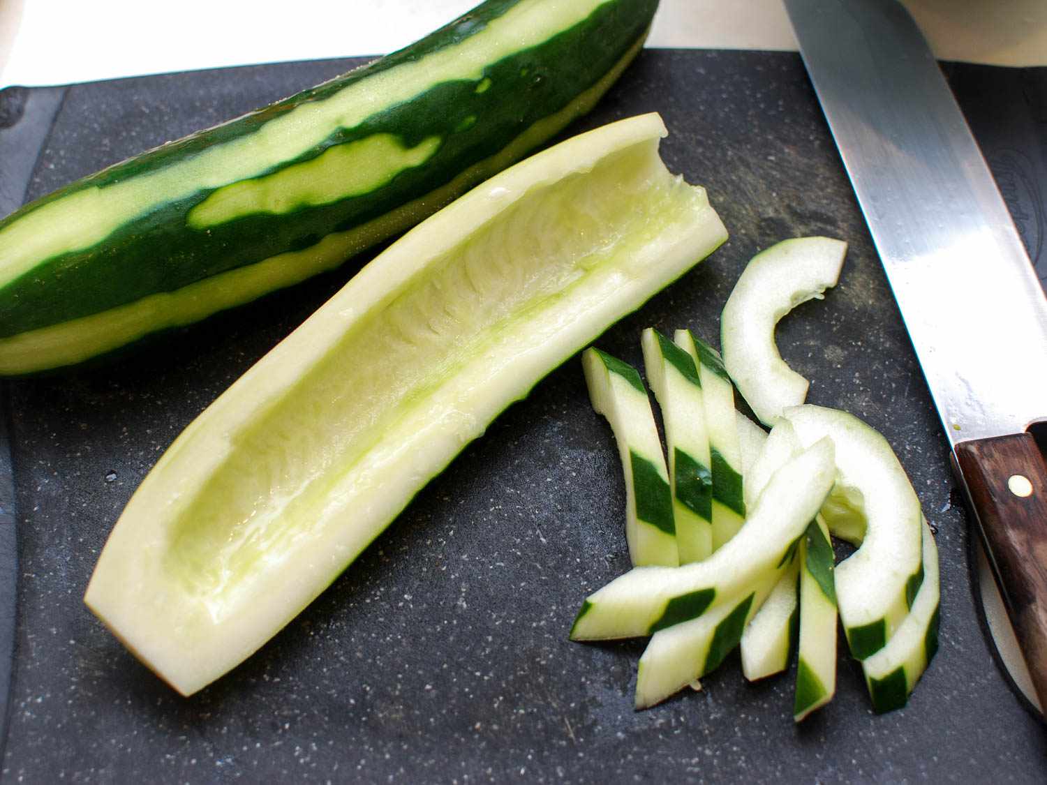 Two cucumbers laying on a cutting board. One is whole with strips of its skin peeled. The other has been halved and deseeded. One half is still intact and the other has been sliced on the bias. The slices have portions of the skin still attached.