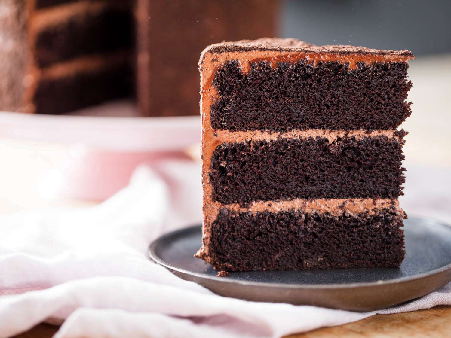 One slice of three-layer chocolate Devil's food cake on a grey plate and pink tablecloth, with remainder of cake in background.