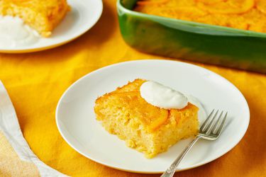 A slice of orange-flavored cake served on a white plate with a dollop of cream and a fork, with the rest of the cake in a green dish in the background