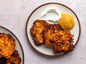 Three crispy sweet potato latkes on a ceramic plate alongside apple sauce and sour cream.