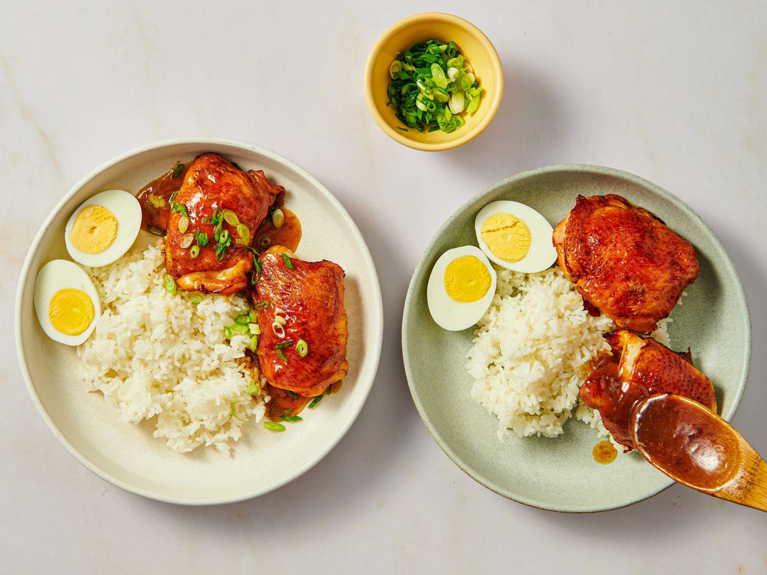 Two plates of a meal featuring roasted chicken rice and boiled eggs with a small bowl of chopped green onions nearby