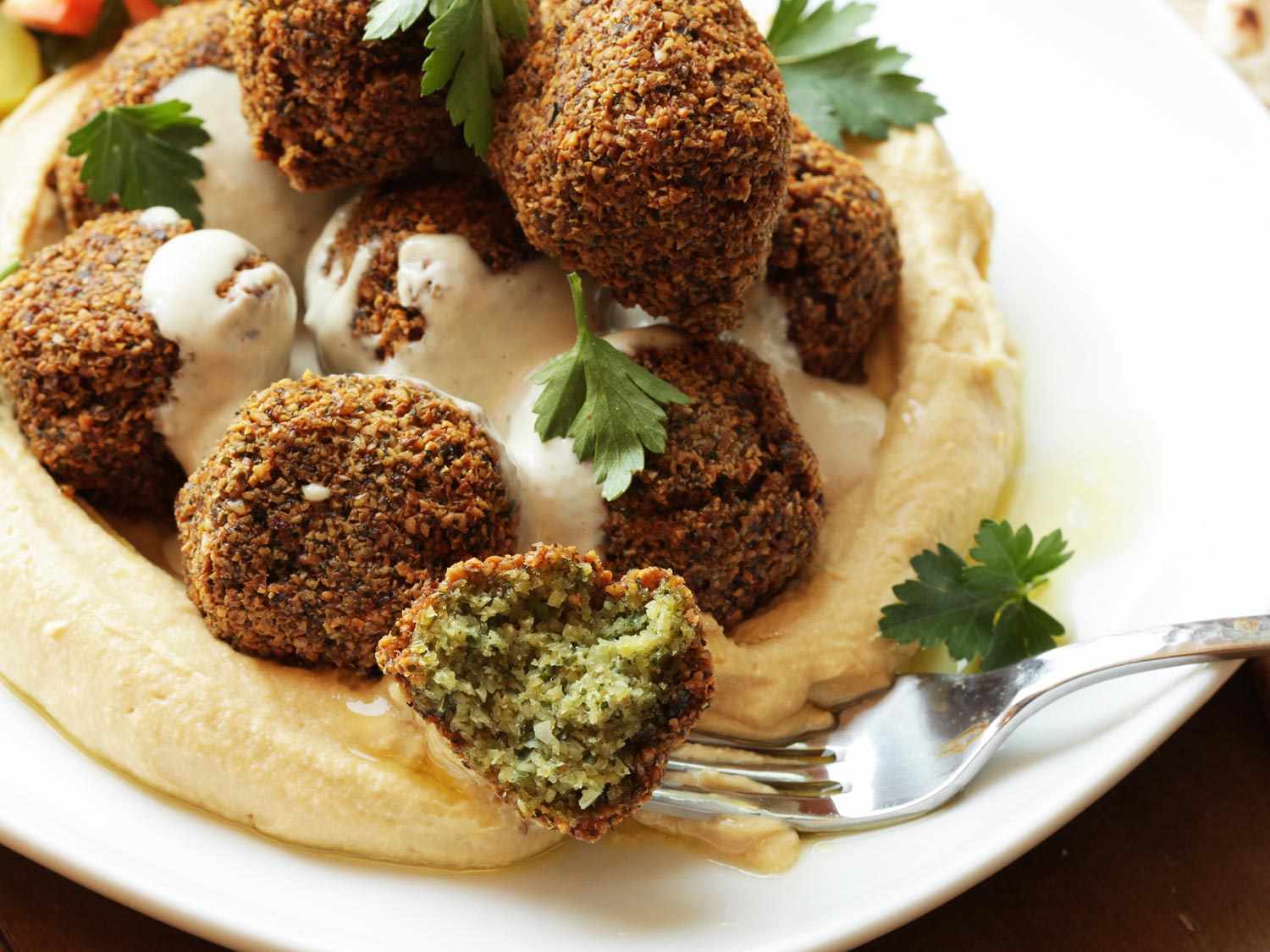 A fork holding part of a falafel ball. There is a plate of more falafel balls and hummus.