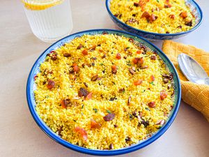 Bowls of couscous garnished with dried fruits and nuts on a table near a drink and a yellow napkin with spoon