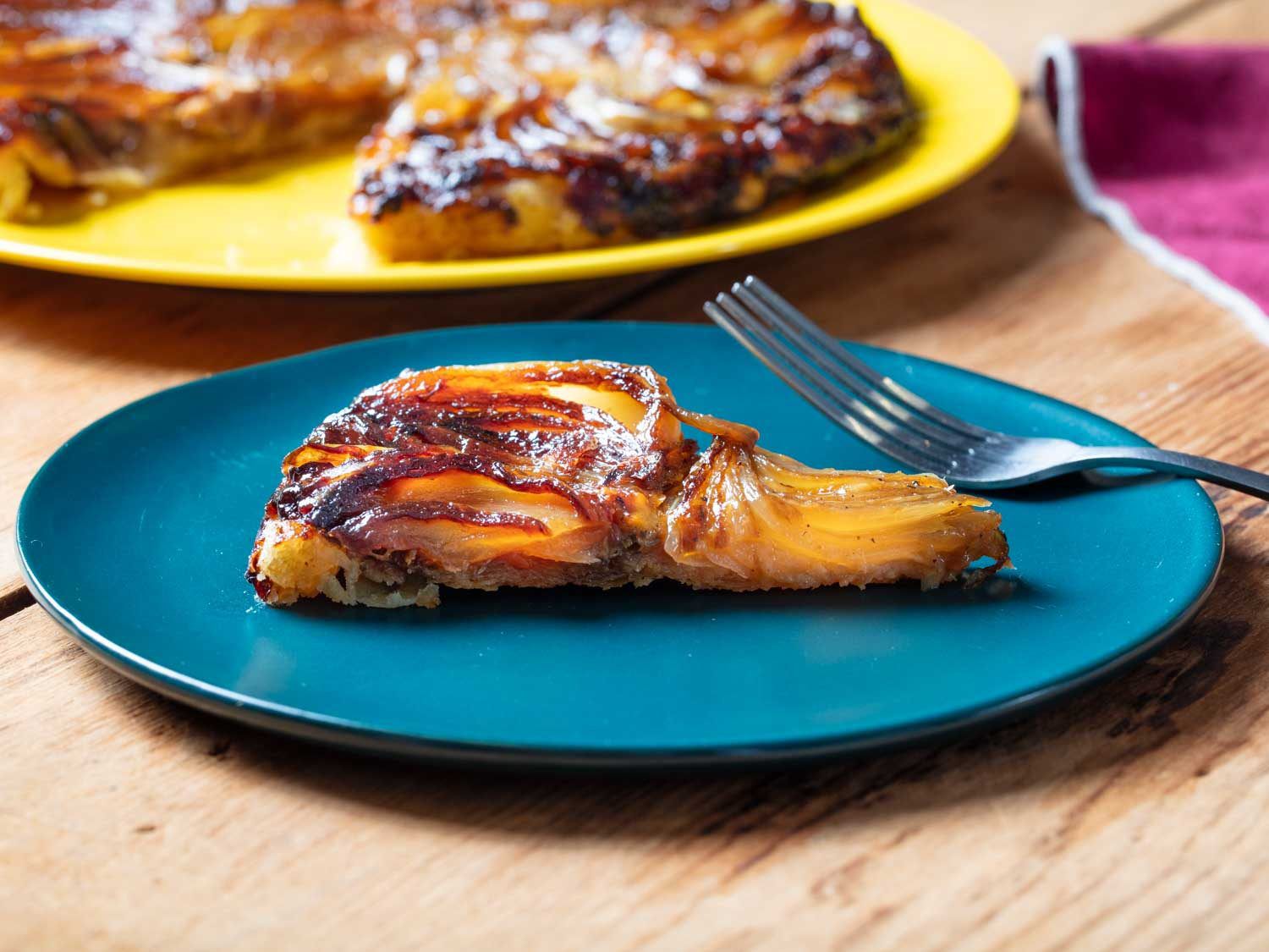 Side view of a slice of tarte tatin on a small plate with the rest of the tarte tatin in the background.