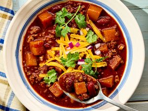 A bowl of turkey chili with squash beans cheese onions and cilantro garnish spoon served alongside