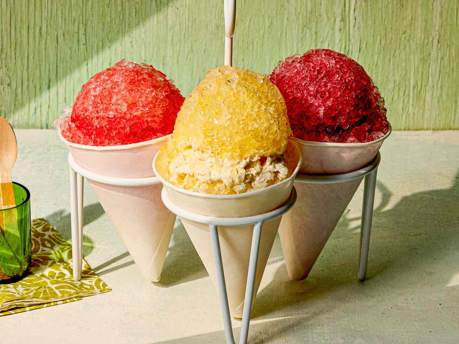 Three shaved ice desserts in cones with red and yellow flavors displayed on cone holders with light green background