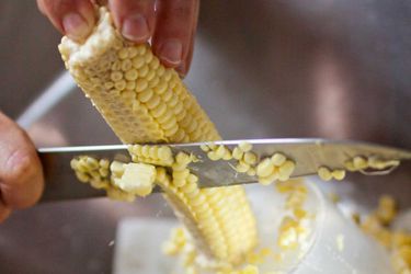 Closeup of hands cutting corn kernels off the cob with a sharp knife.