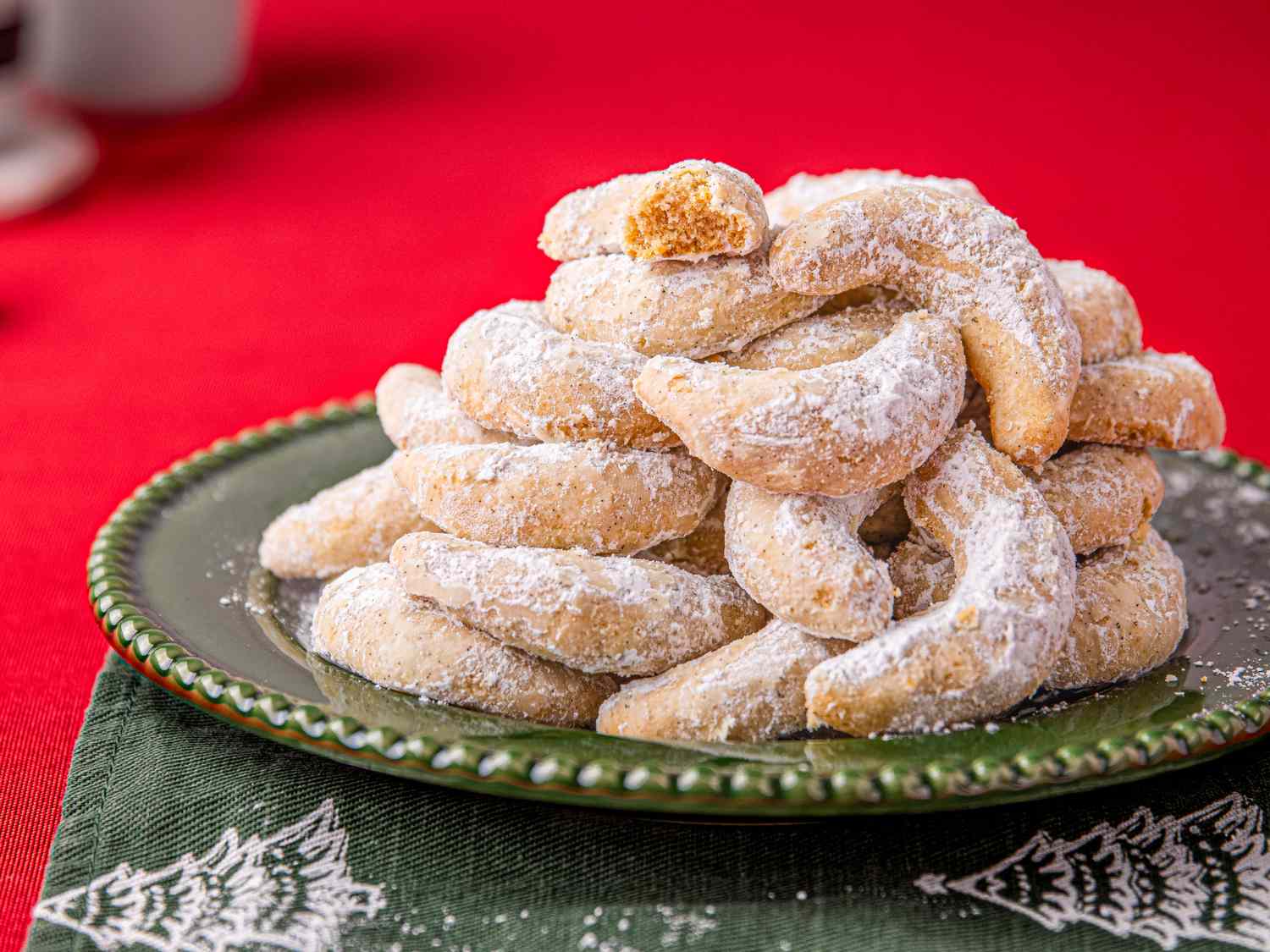 A stack of crescentshaped cookies dusted with powdered sugar arranged on a green plate placed over a festive napkin