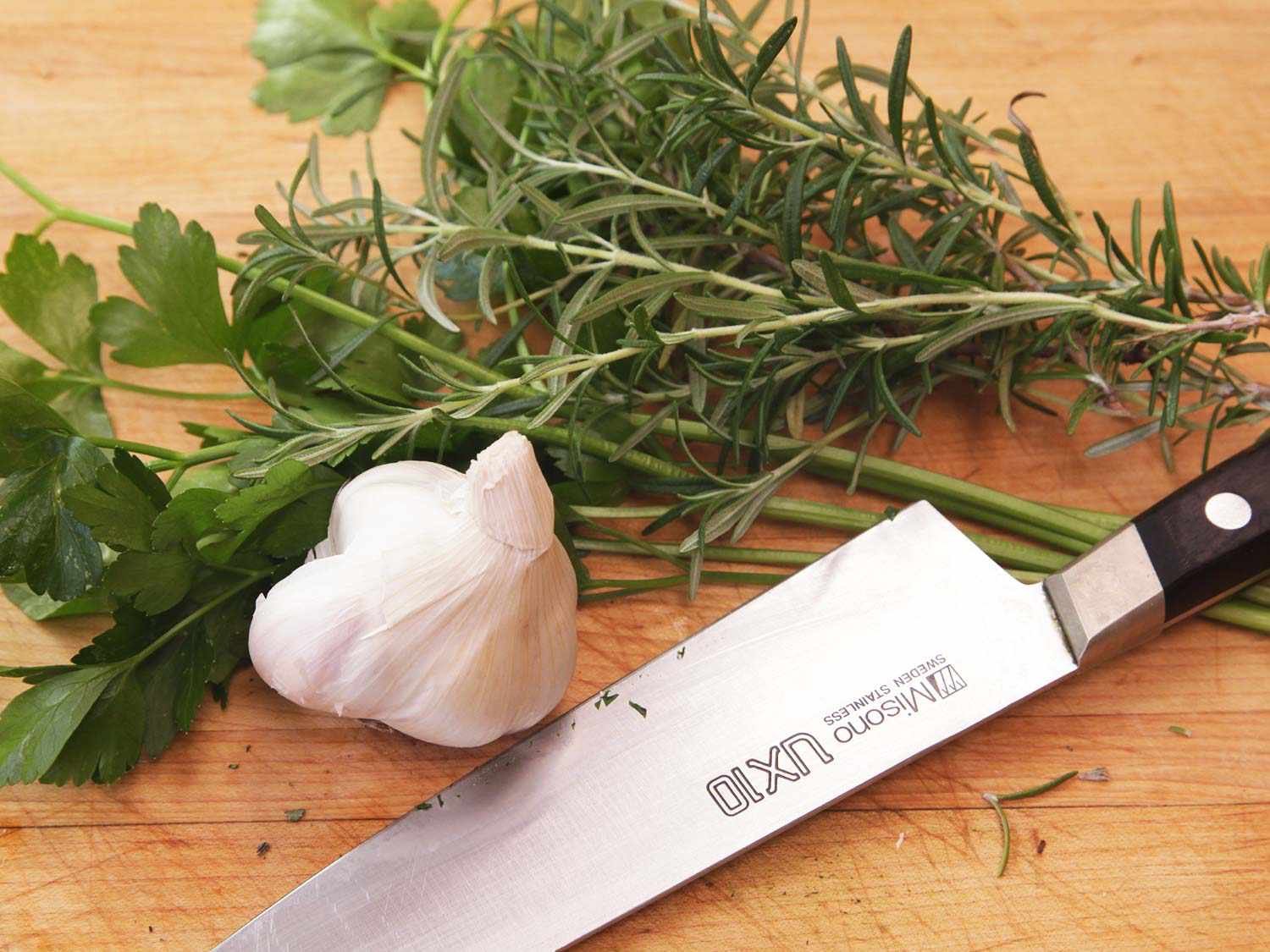 Parsely, rosemary, and garlic resting on a cutting board alongside a knife.
