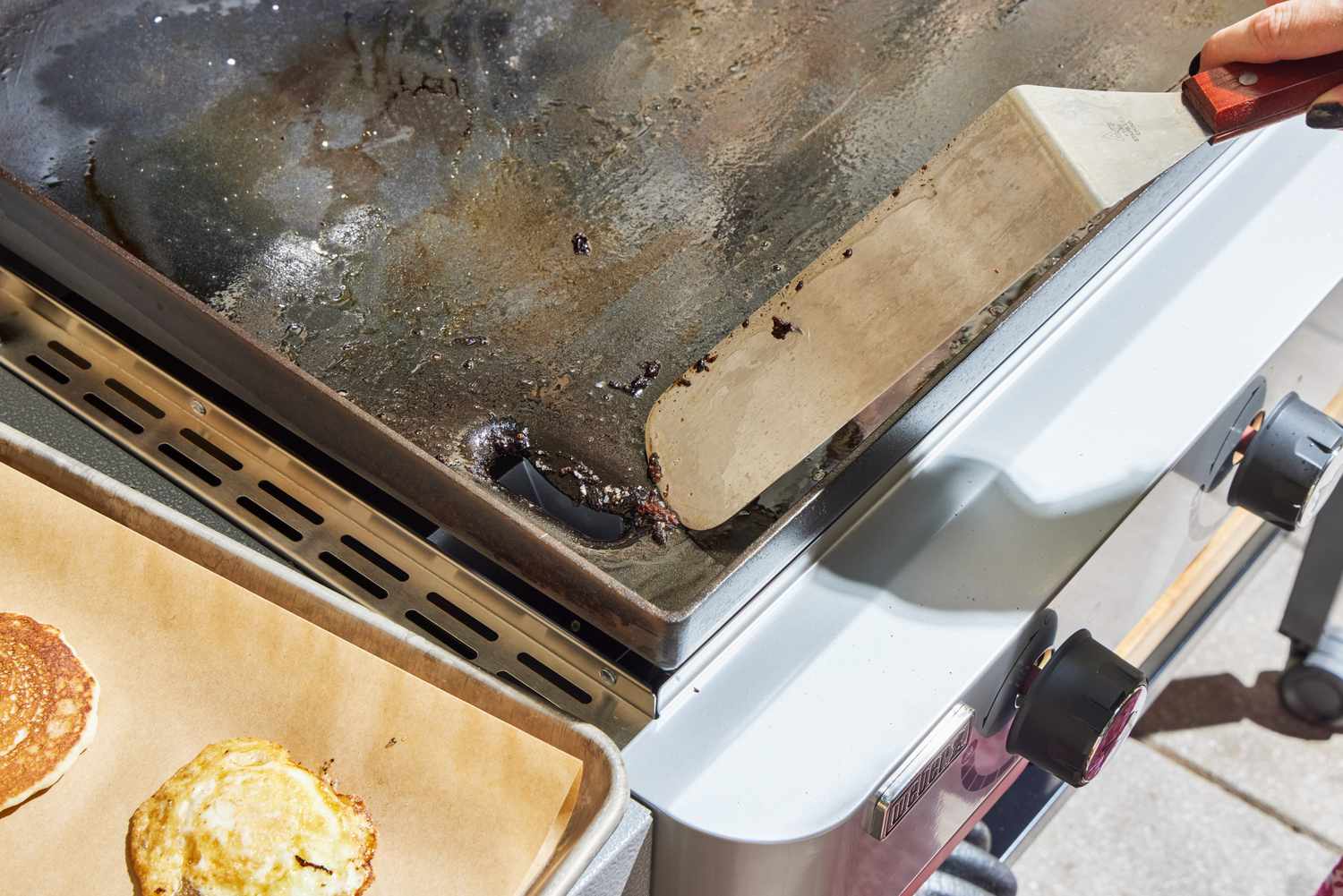 A person cleans the Weber 36 Inch Gas Griddle using a flat spatula