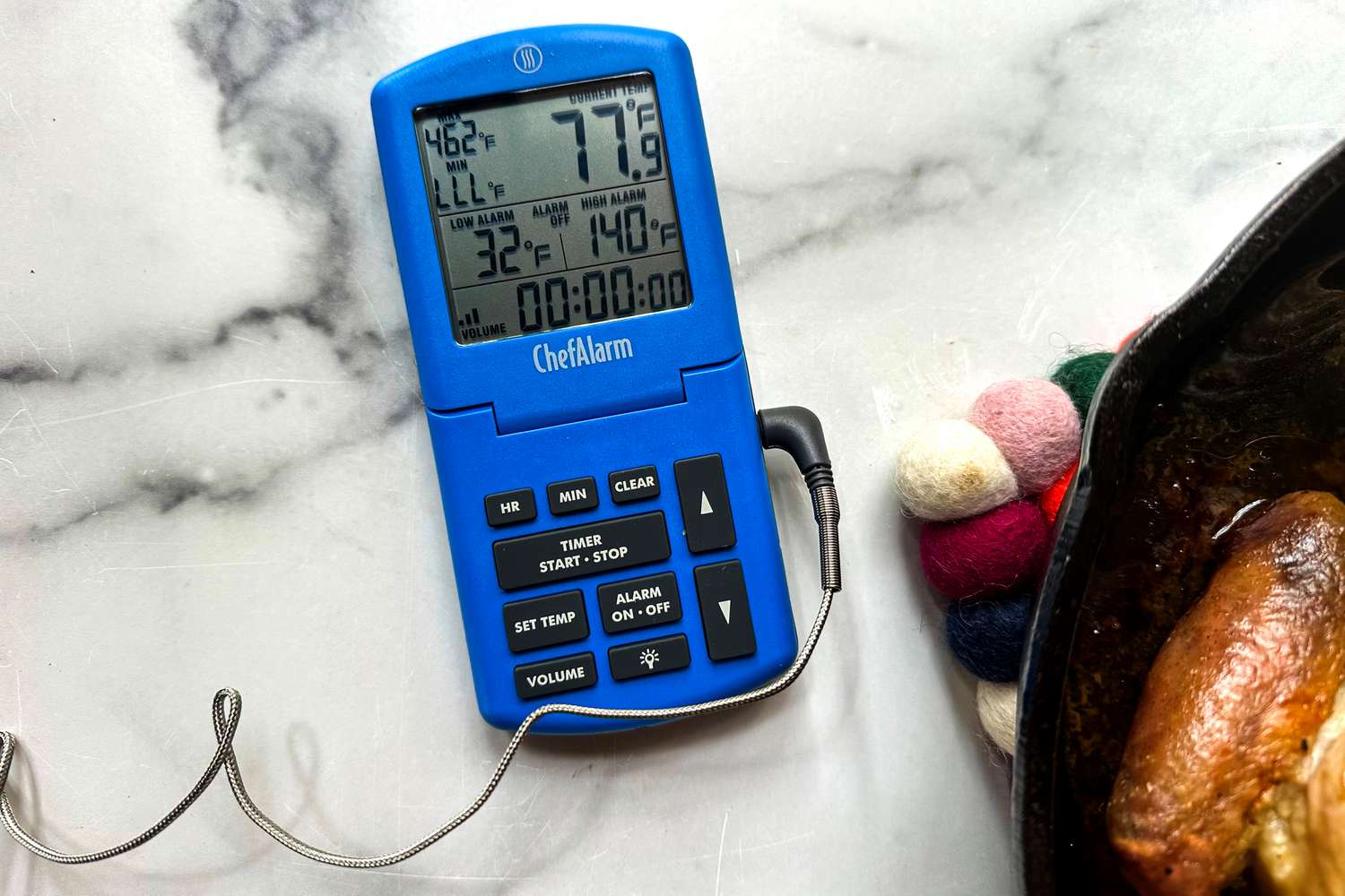 A blue digital oven thermometer on a marble surface next to a cast iron pan.