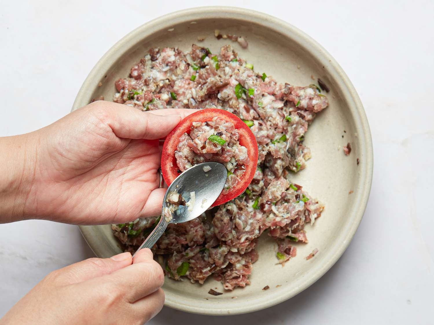 A hand spooning minced pork mixture into a halved tomato resting over a bowl of the same mixture