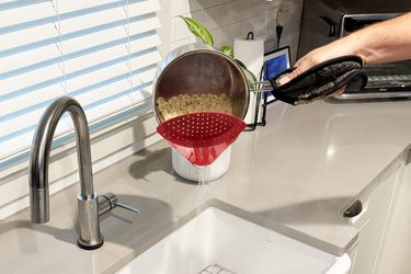 A person using a clip-on strainer to water from pasta noodles into a sink.