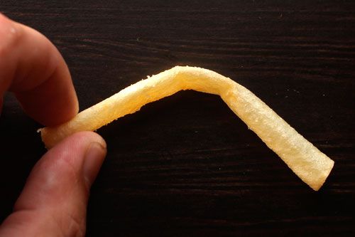 A closeup of fingers holding a french fry that bends in half at the middle against a black background.