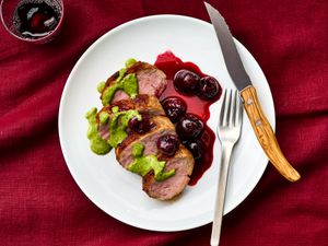 Sliced roasted pork tenderloin with green sauce and roasted cherries on a plate, placed on a red tablecloth with a fork and knife