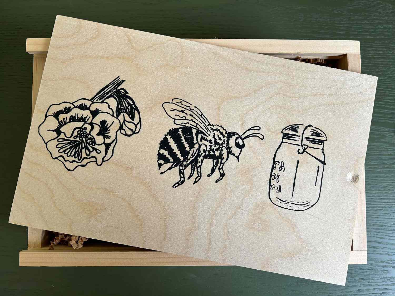 A wooden box with etched drawings of a flower, bee, and jar