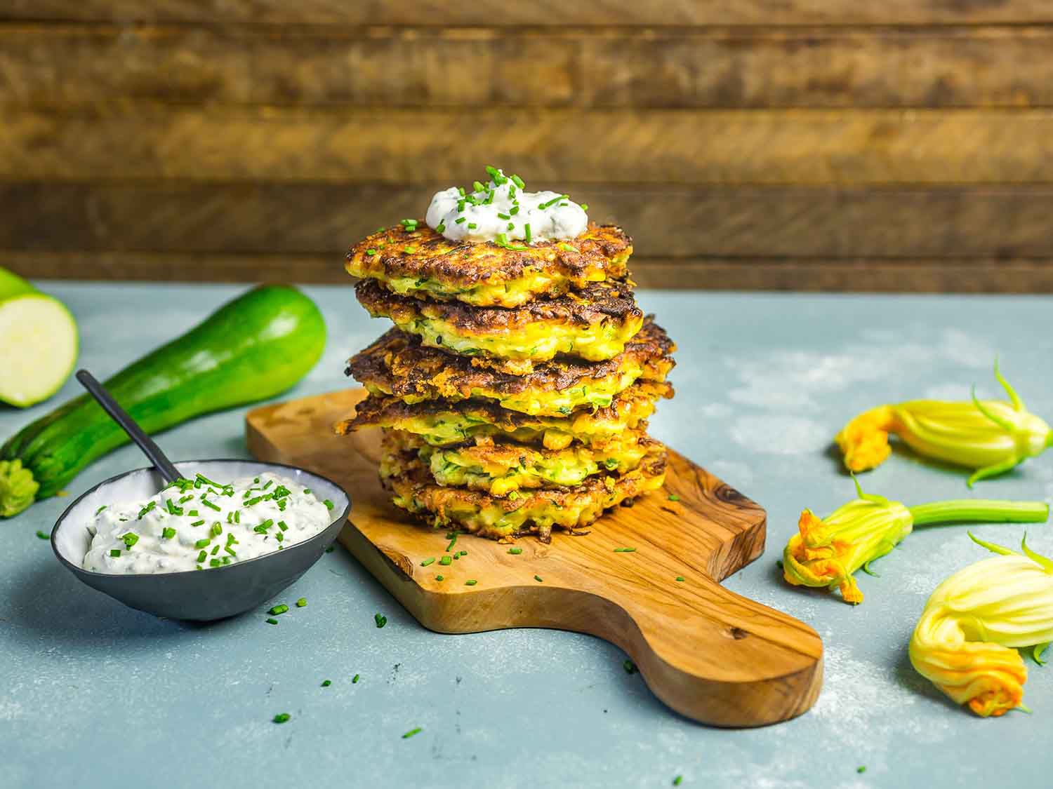 A stack of zucchini-and-corn fritters on a wooden board, topped with herb sour cream, surrounded by a bowl of sauce, squash, and squash blossoms.