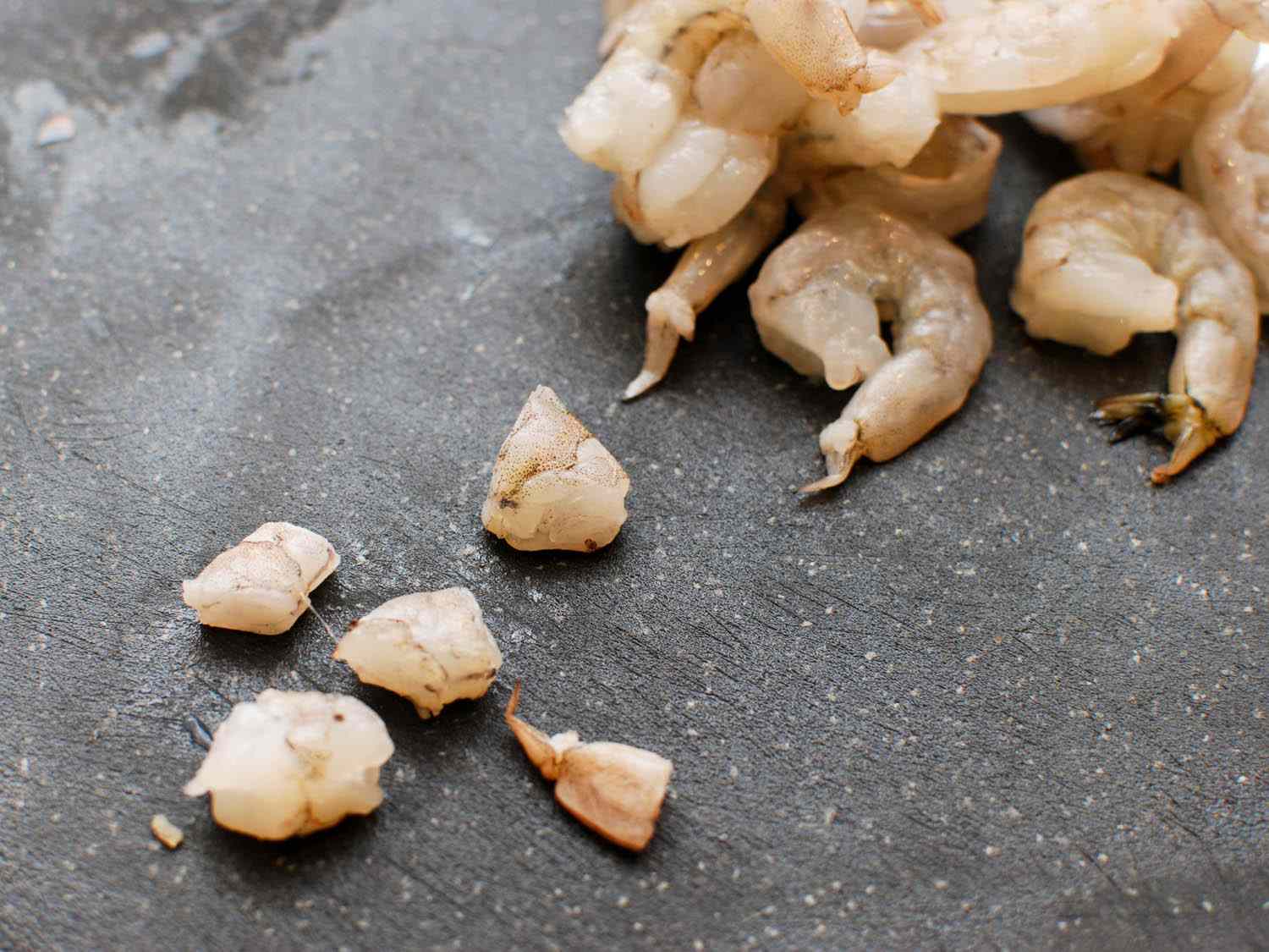 A raw, marinated shrimp on a cutting board, chopped into 5 pieces. A pile of whole, peeled shrimp are off in the right upper corner.