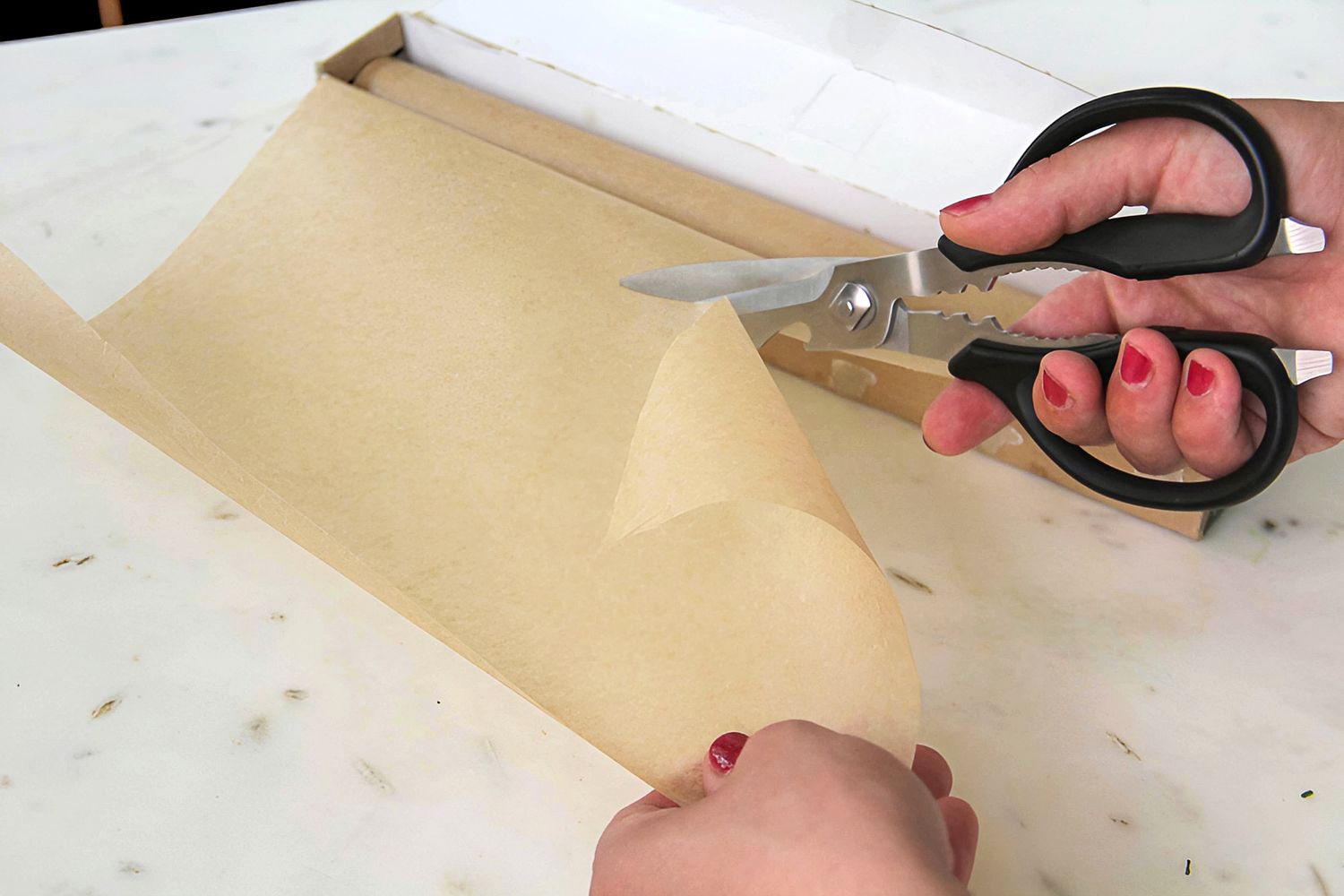 Hands using Cangshan 9-inch Heavy-Duty Come-Apart Kitchen Shears to cut nonstick parchment paper from a roll on a white surface